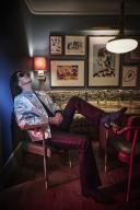 Per dressed in black pants and a silver jacket reclining in a chair.