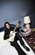 Per in a black suit with a white electric guitar on a bed.